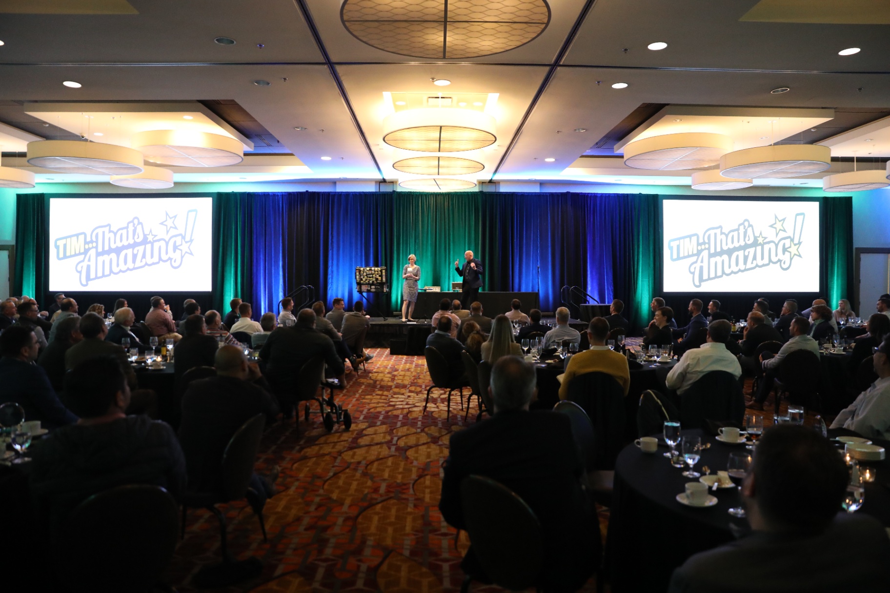 Wide shot of Tim on stage in a hotel ballroom with a corporate audience