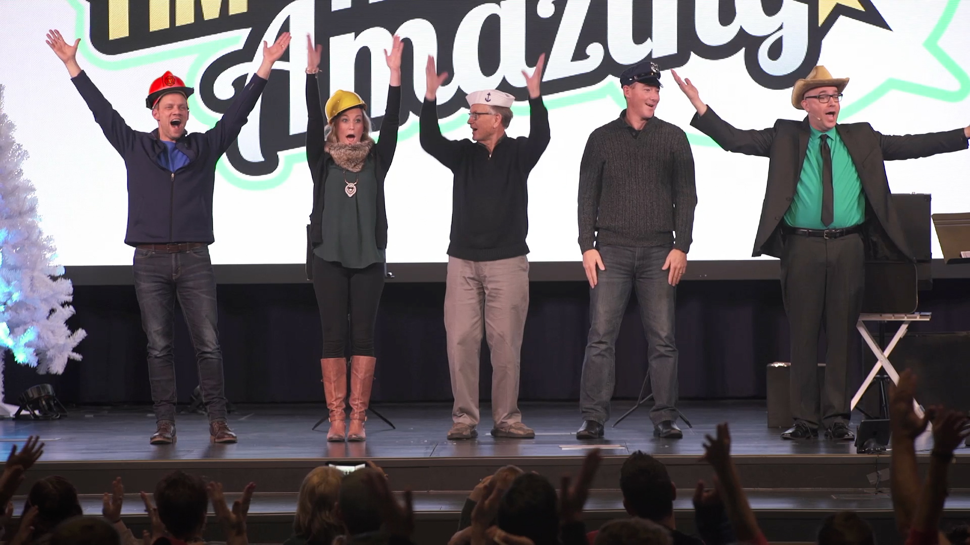 Five volunteers on stage with Tim wearing costume hats, arms raised